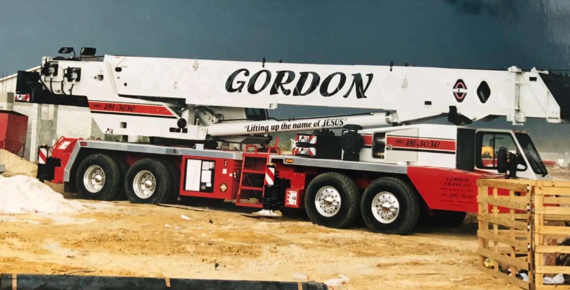 Gordon Crane Co.'s red and white truck crane parked at a sandy jobsite under a dark sky, GORDON lettered in black script along the white boom and “Lifting up the name of JESUS” painted down the side.